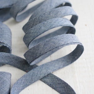 Könnte beinhalten: Ein durchgehendes Stück blaues Chambray-Schrägband, das zu einer Spirale gewickelt ist. Der Stoff weist eine gewebte Textur mit einer dezent melierten Optik auf und zeigt eine gleichmäßige Mischung aus blauen und weißen Fäden.