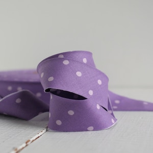 May include: A roll of purple fabric ribbon featuring a repeating pattern of small, light pink polka dots. The ribbon is coiled in a loose, overlapping loop, displaying a smooth, woven texture throughout the length of the material.