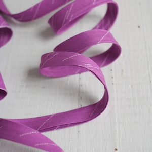 Könnte beinhalten: Ein Band aus leuchtend violettem Schrägband mit einem dezenten weißen diagonalen Streifenmuster. Der Stoff ist gefaltet und gebogen, wodurch ein fließendes, bandartiges Aussehen entsteht. Ideal für Näh- und Bastelprojekte.