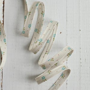 May include: A length of off-white bias tape featuring a repeating pattern of small light blue flowers with green leaves and tiny brown stems. The fabric trim is folded into a continuous strip, displaying a consistent floral print throughout the length.