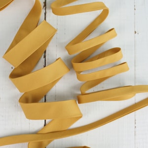 May include: A length of mustard yellow fabric ribbon, folded and arranged on a white wooden surface.