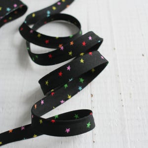 May include: Black ribbon with a repeating pattern of colorful stars in red, yellow, green, and pink. The ribbon is coiled and laid out on a white wooden surface. A roll of the same ribbon is visible in the background.