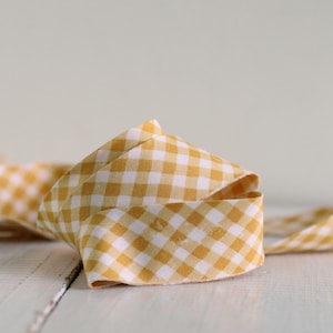 May include: A length of fabric ribbon featuring a yellow and white gingham check pattern. The ribbon is loosely coiled, showing the woven texture of the material and the consistent repeating square pattern across the entire surface of the textile.