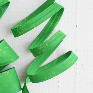 Puede incluir: Una tira de cinta al bies de color verde brillante doblada en forma de espiral. La tela presenta un patrón moteado con pequeños puntos dispersos en amarillo y azul claro por toda la superficie del material verde, creando un aspecto texturizado.