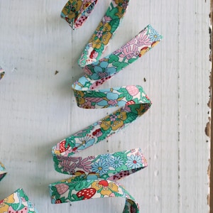 May include: A length of fabric ribbon featuring a repeating pattern of colorful flowers, green leaves, and red strawberries on a light blue background. The ribbon is coiled in a spiral shape, displaying the intricate floral and fruit print design.
