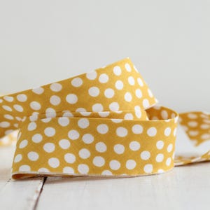 May include: A mustard yellow fabric ribbon featuring a repeating pattern of small white polka dots. The ribbon is coiled in a loose, overlapping loop, displaying the woven texture of the material and the consistent spacing of the circular print.
