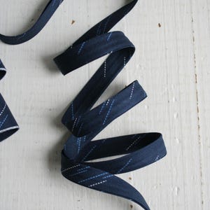 Könnte beinhalten: Ein Band in Marineblau mit einem dezenten Muster aus weißen und hellblauen diagonalen Linien. Das Band ist gewellt und liegt auf einer weißen, strukturierten Oberfläche, wodurch seine flexible Form und sein Design zur Geltung kommen.