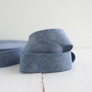 Può includere: Un rotolo di nastro in tessuto denim blu con una trama testurizzata e un bordo leggermente sfilacciato. Il nastro è avvolto in una spirale allentata, mostrando il colore blu medio e il visibile motivo incrociato del materiale tessile.