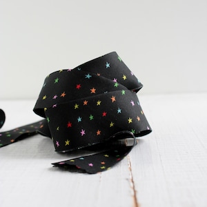Könnte beinhalten: Schwarzes Stoffband mit einem Muster aus bunten Sternen. Das Band ist gerollt und zeigt das Sternmuster in Rot, Gelb, Grün, Blau und Rosa. Es liegt auf einer weißen Holzoberfläche, die einen Kontrast bildet.