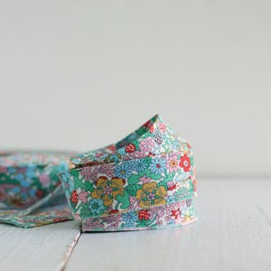 May include: A coiled length of fabric ribbon featuring a dense, colorful floral pattern. The design includes small red strawberries, yellow flowers, light blue daisies, and green leaves on a white background, with a visible woven texture throughout.