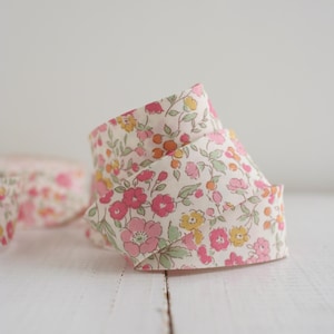 May include: A roll of floral ribbon with a cream background and pink, yellow, and green flowers. The ribbon is curled, showing the pattern and texture. Another roll of ribbon is visible in the background.