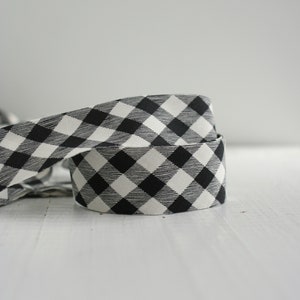 May include: A roll of fabric ribbon featuring a black and white gingham check pattern. The ribbon is coiled, showing the repeating square pattern of alternating black and white colors across the woven textile surface of the material.