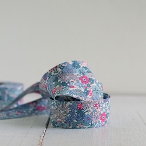 May include: A roll of fabric ribbon featuring a dense floral pattern in shades of blue, light pink, and white. The ribbon is coiled, showing a repeating design of small flowers, leaves, and stems across the entire surface of the textile material.