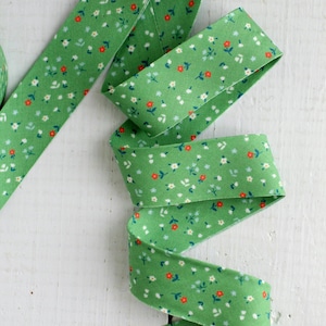 Puede incluir: Una tira larga de cinta de tela verde con un patrón repetitivo de diminutas flores blancas y rojas con pequeños tallos verdes. La cinta está doblada en forma de zigzag, mostrando claramente el estampado floral a lo largo de todo el material.