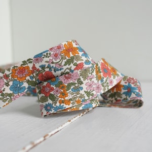 Peut inclure: Ruban floral sur fond blanc. Le ruban présente un motif répétitif de fleurs colorées dans les tons orange, rose, bleu et vert. Le ruban est plié sur lui-même, créant un effet de superposition.