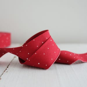 May include: A length of red fabric ribbon featuring a repeating pattern of small white polka dots. The ribbon is coiled in a loose loop, displaying the vibrant red color and the consistent distribution of the white dots across the textile surface.
