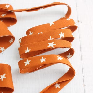 Könnte beinhalten: Nahaufnahme eines orangefarbenen Stoffschrägbandes mit einem sich wiederholenden Muster aus weißen Sternen. Das Band ist gefaltet und gebogen, wodurch ein strukturierter Effekt entsteht. Der Stoff ist wahrscheinlich Baumwolle, geeignet für Näh- und Bastelprojekte.