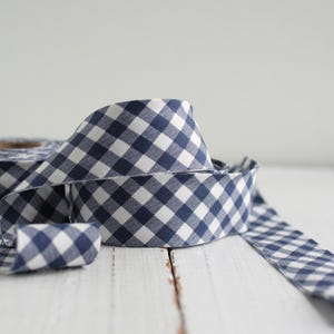 Puede incluir: Un rollo de cinta vichy azul y blanca. La cinta está doblada y colocada sobre una superficie de madera blanca.