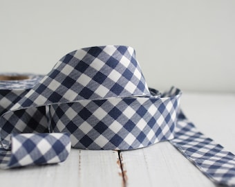 May include: A roll of fabric ribbon featuring a classic navy blue and white gingham check pattern. The ribbon is coiled, with one end extending outwards, displaying the consistent diagonal grid design across the woven textile surface.