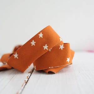 Op de afbeelding: Oranje stoffen lint met een herhalend patroon van witte sterren. Het lint is over zichzelf gevouwen, waardoor een gelaagd effect ontstaat. Het lint ligt op een wit houten oppervlak.