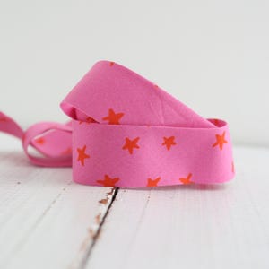 May include: A coiled pink ribbon featuring a repeating pattern of orange stars. The ribbon is placed on a white wooden surface. The ribbon is made of fabric.