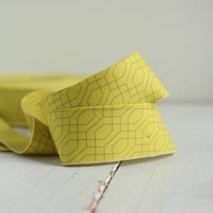 May include: A roll of yellow fabric ribbon featuring a repeating dark gray geometric pattern of interconnected octagons and lines. The ribbon is folded over itself, showing the textured weave of the material and the consistent printed design.