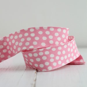 May include: A coiled length of fabric ribbon featuring a bright pink background with a repeating pattern of small, solid white polka dots. The ribbon is made of a woven textile material and is displayed in a loose, overlapping spiral formation.