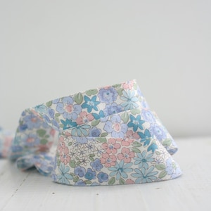 Pode incluir: Uma fita de tecido com um padrão floral repetitivo de flores azul-claras, rosa suave e verde-sálvia sobre um fundo branco. A fita está enrolada numa espiral solta, exibindo a intrincada estampa botânica em toda a sua extensão.