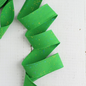Puede incluir: Una cinta al bies de color verde vibrante con un patrón moteado de pequeños puntos amarillos y azules. El ribete de tela está doblado en una forma continua y sinuosa, mostrando el color uniforme y el diseño de salpicaduras de pintura.
