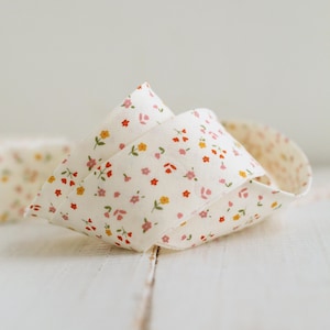 Puede incluir: Una cinta de tela de color blanco roto enrollada con un patrón repetitivo de diminutas y delicadas flores en tonos rosa, naranja y amarillo, con pequeños tallos y hojas verdes, dispuesta en un bucle suelto y plegado sobre una superficie.