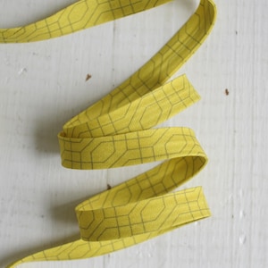 Könnte beinhalten: Ein gelbes Stoffband mit einem geometrischen Muster in einem dunkleren Gelbton. Das Band ist gewellt und liegt auf einer weißen Oberfläche. Die Breite des Bandes beträgt ca. 2 cm.