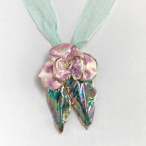 Puede incluir: Un collar con colgante y cinta verde claro. El colgante presenta un diseño floral rosa y blanco con detalles dorados y hojas verdes iridiscentes. El collar se exhibe en un maniquí blanco.