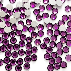 May include: A large quantity of small, round, purple rhinestones with silver backs. The rhinestones are scattered on a white surface.