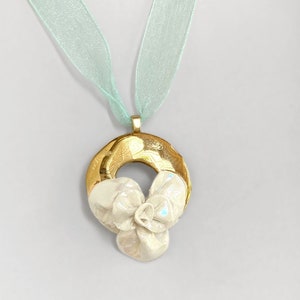 Puede incluir: Un collar con un colgante de cerámica dorado y blanco con diseño de flor. El collar está en una cinta azul claro.