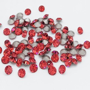 Puede incluir: Una dispersión de piedras de cristal rojas con respaldos plateados. Las piedras de cristal son pequeñas y redondas, y están dispersas sobre un fondo blanco.