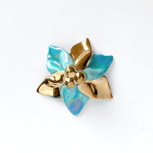 Puede incluir: Una flor de cerámica azul y dorada con forma de estrella de cinco puntas. La flor tiene un acabado brillante y tres bolas doradas en el centro.