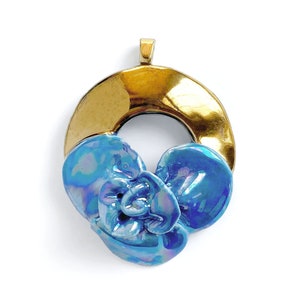 Puede incluir: Un colgante dorado y azul con un diseño de flor. El colgante está hecho de metal y tiene un acabado brillante.