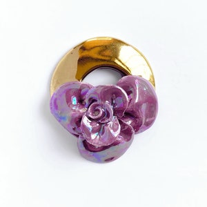 Puede incluir: Una flor de cerámica morada con detalles iridiscentes está unida a un anillo dorado.