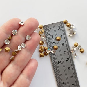 May include: A close-up of a hand holding a variety of gold and clear rhinestones. A ruler is visible in the background, with measurements in inches and centimeters.