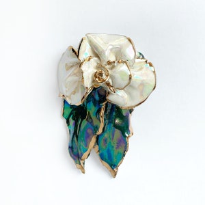 Puede incluir: Un broche de cerámica blanca en forma de flor con hojas verde y azul iridiscentes. La flor tiene un centro dorado y está adornada con un borde dorado.
