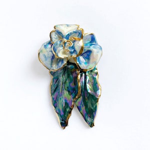 Puede incluir: Un broche de rosa de porcelana azul y blanco con hojas verdes iridiscentes y detalles dorados.