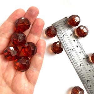 May include: A collection of round, faceted, translucent amber-colored beads. A metal ruler is positioned next to the beads, showing each bead has a diameter of approximately 0.5 inches. The beads feature a central hole for threading.