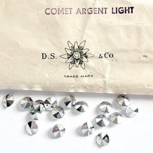 Peut inclure: Un ancien paquet de strass "COMET ARGENT LIGHT" avec le texte "D.S. & Co." et "TRADE MARK". L'emballage est beige et légèrement froissé, avec une dispersion de strass argentés en dessous.