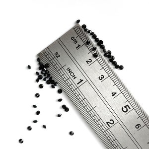 Puede incluir: Cristales negros esparcidos sobre una superficie blanca junto a una regla de metal marcada en pulgadas y cent&iacute;metros. La regla muestra medidas de hasta 6 pulgadas o 15 cent&iacute;metros.