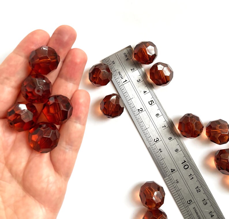 May include: A collection of round, faceted, translucent amber-colored beads. A metal ruler is positioned next to the beads, showing each bead has a diameter of approximately 0.5 inches. The beads feature a central hole for threading.