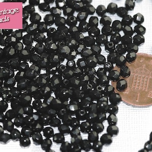 May include: A close-up image of a large quantity of black faceted glass beads. The beads are approximately the size of a British one pence coin, which is shown in the image for scale.
