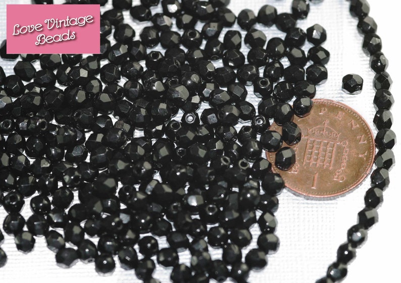 May include: A close-up image of a large quantity of black faceted glass beads. The beads are approximately the size of a British one pence coin, which is shown in the image for scale.