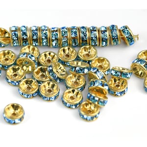 May include: A group of gold-toned bead caps with a single row of light blue rhinestones around the edge. The bead caps are scattered on a white surface.