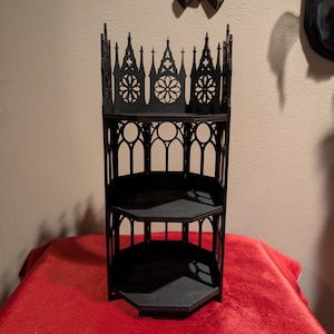 May include: A dark brown, three-tiered shelf with a gothic architectural design. The shelf has an open back and features decorative cutouts, including arched windows and floral patterns. It is displayed on a red surface.