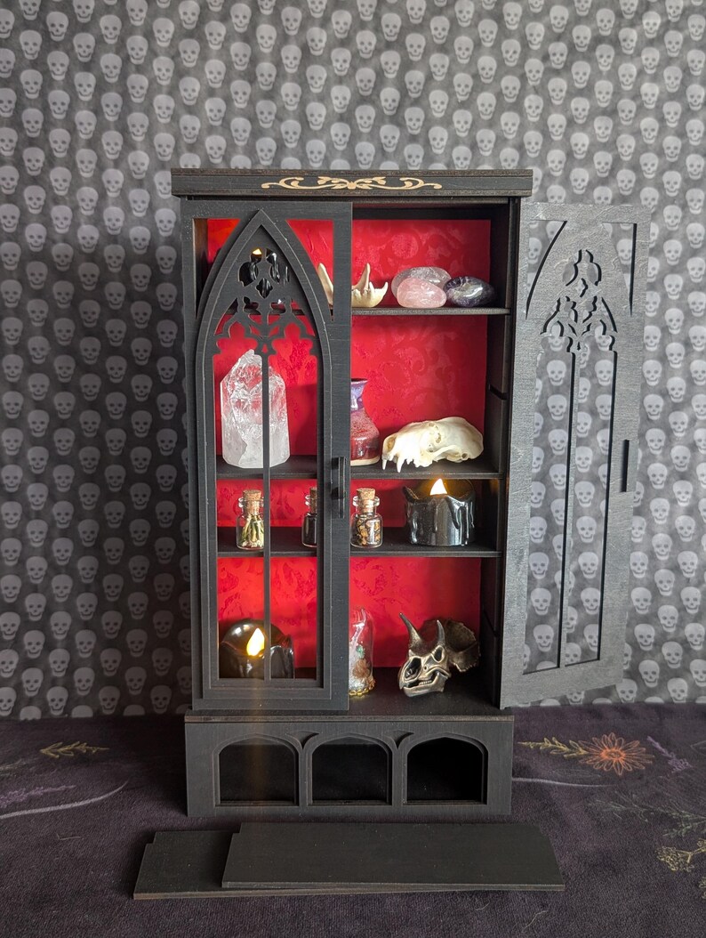 K&ouml;nnte beinhalten: Ein schwarzer Kuriosit&auml;tenschrank im gotischen Stil mit rotem Innenraum, der verschiedene Gegenst&auml;nde ausstellt. Der Schrank hat eine Schiebet&uuml;r mit Bogenmuster. Zu den Artikeln geh&ouml;ren Kristalle, ein Sch&auml;del und kleine Flaschen. Der Hintergrund ist ein graues Sch&auml;delmuster.